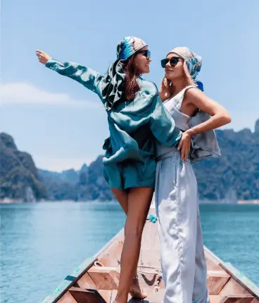 Dos amigas en bote, con pañuelos y gafas de sol, disfrutando de un lago con montañas.